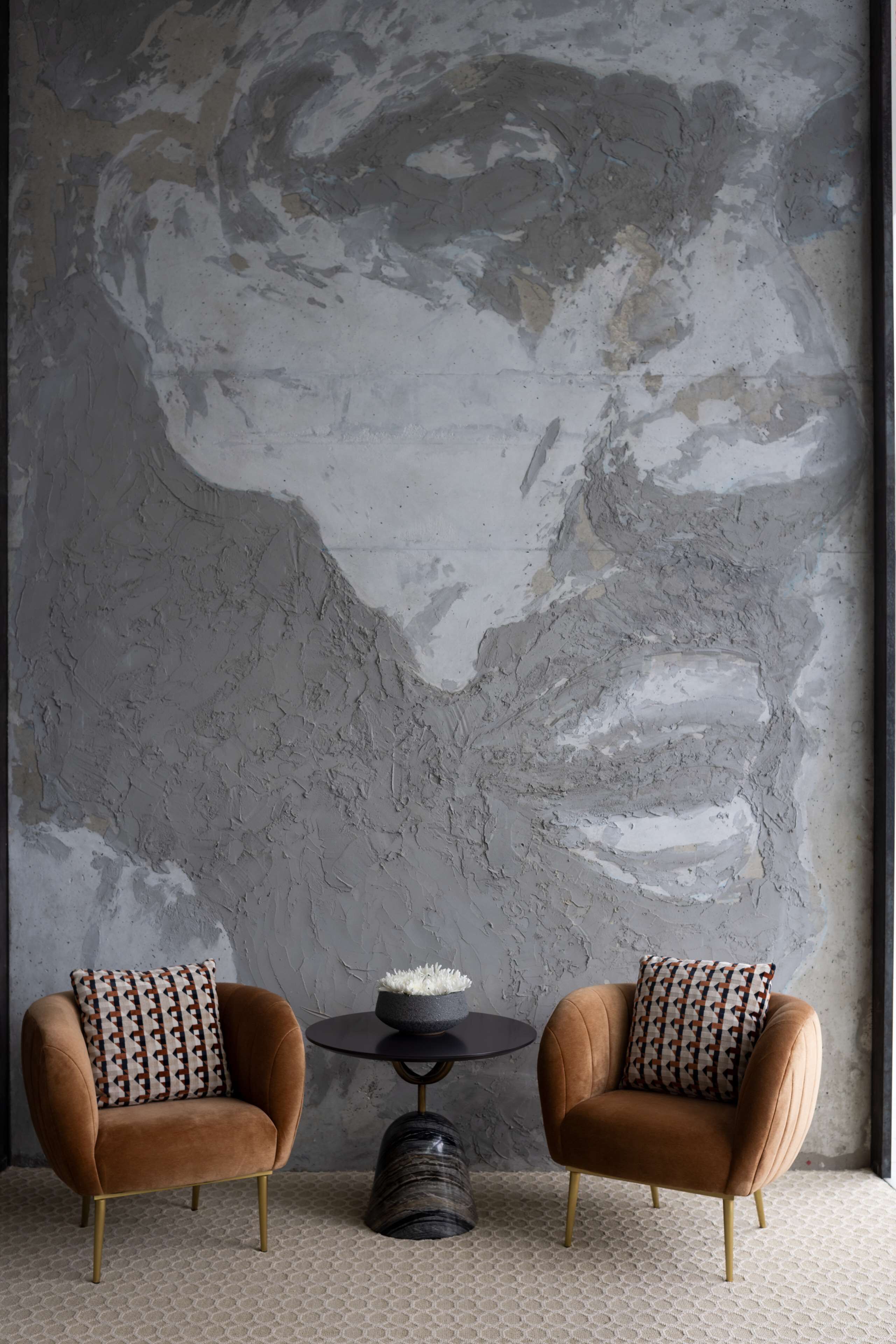 A modern room with two brown armchairs, a round table with a white fluffy pillow, and a textured gray wall.