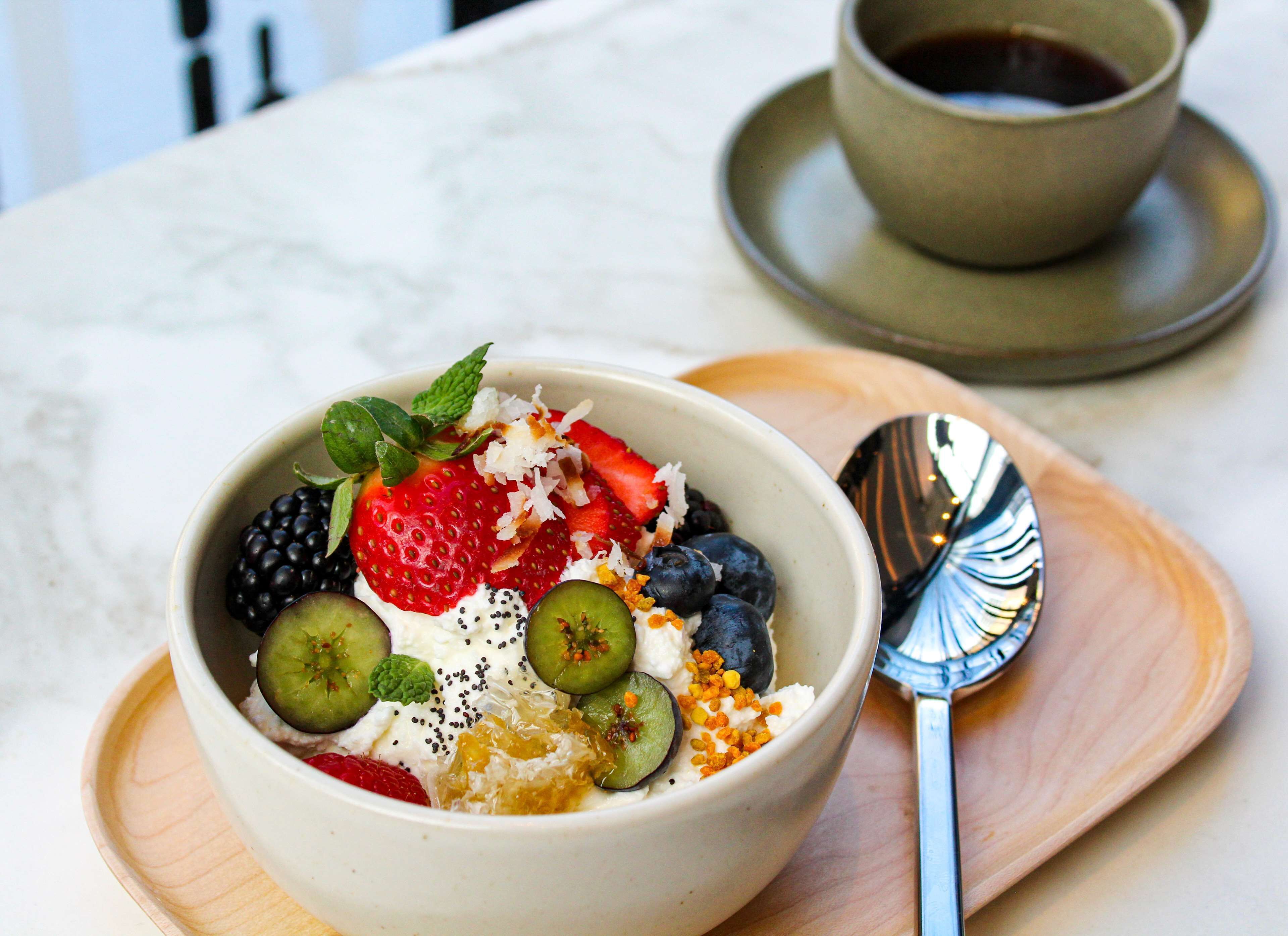 A bowl of fruit salad with yogurt and seeds, a spoon, and a cup of coffee on a wooden tray.