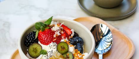A bowl of fruit salad with yogurt and seeds, a spoon, and a cup of coffee on a wooden tray.