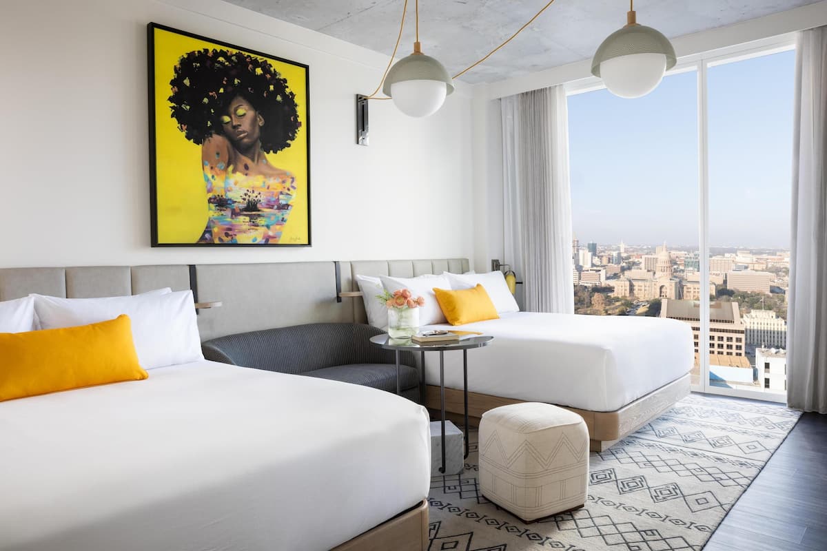 A hotel room with two beds, a small table with a vase of flowers, and a view of the city through large windows.