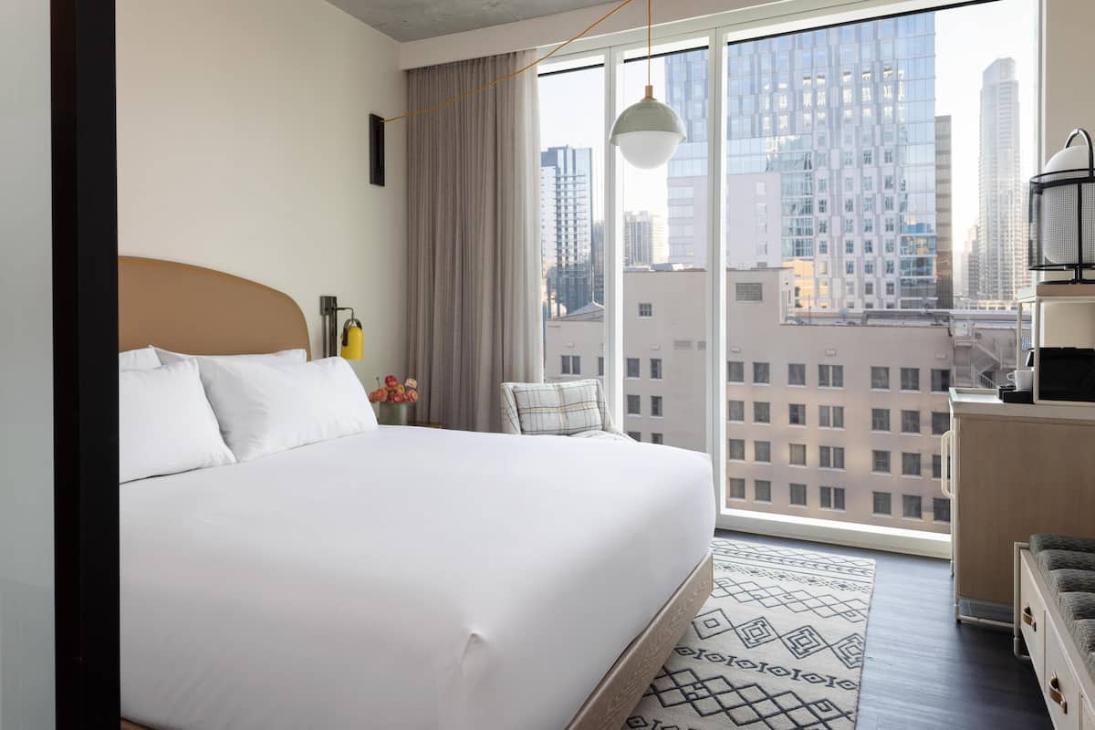 A modern hotel room with a large bed, a view of the city through floor-to-ceiling windows, a small desk with a lamp, and a cityscape view.