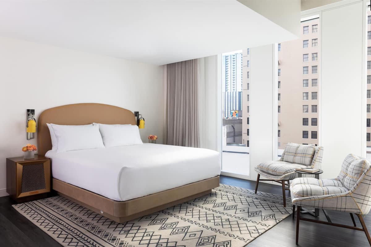 A hotel room with a large bed, a nightstand with a lamp, a chair, and a view of the city through the window.