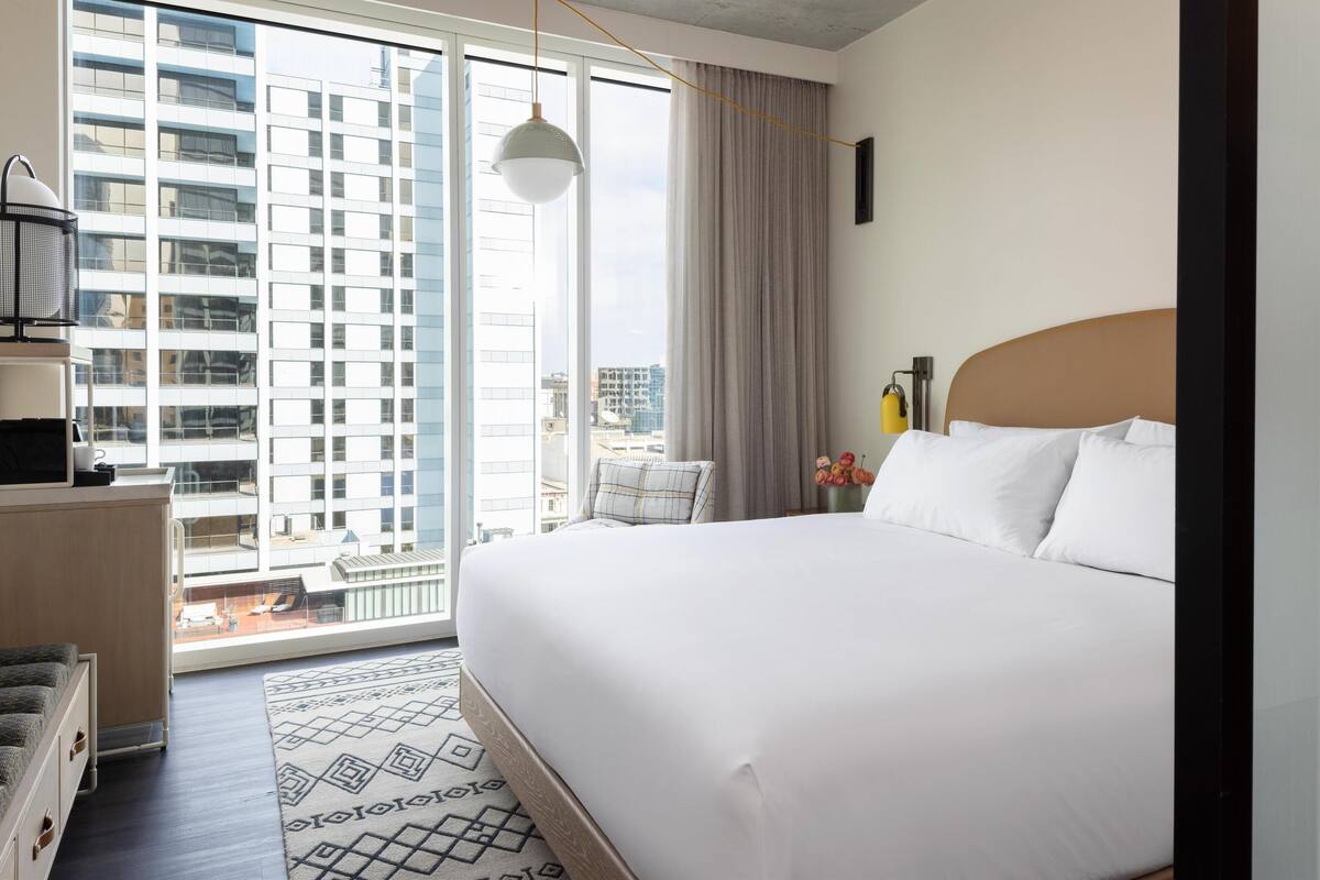 A hotel room with a large bed, a view of a cityscape through floor-to-ceiling windows, a small seating area, and a modern pendant light.
