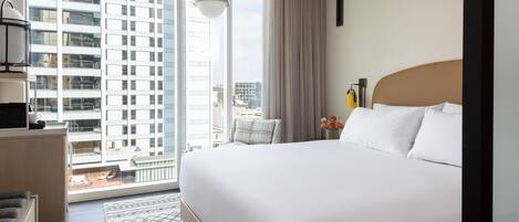 A hotel room with a large bed, a view of a cityscape through floor-to-ceiling windows, a small seating area, and a modern pendant light.