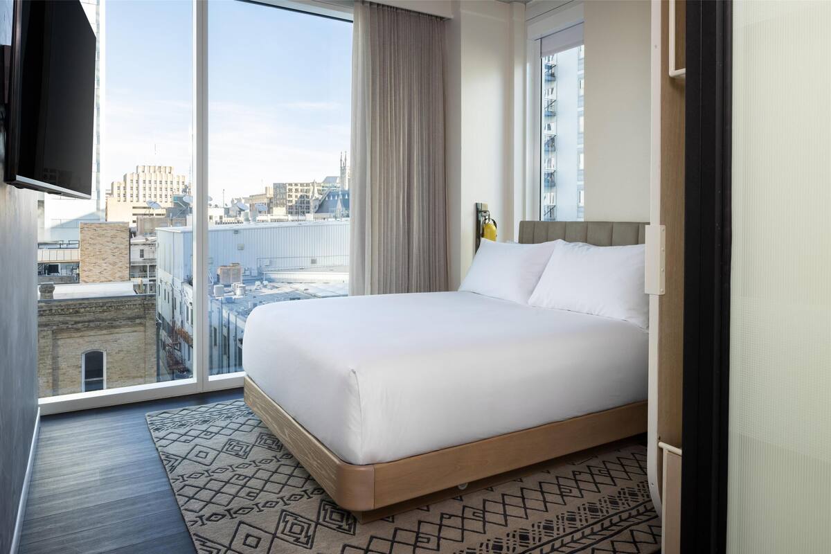 A hotel room with a large bed, a view of the city, and a TV mounted on the wall.
