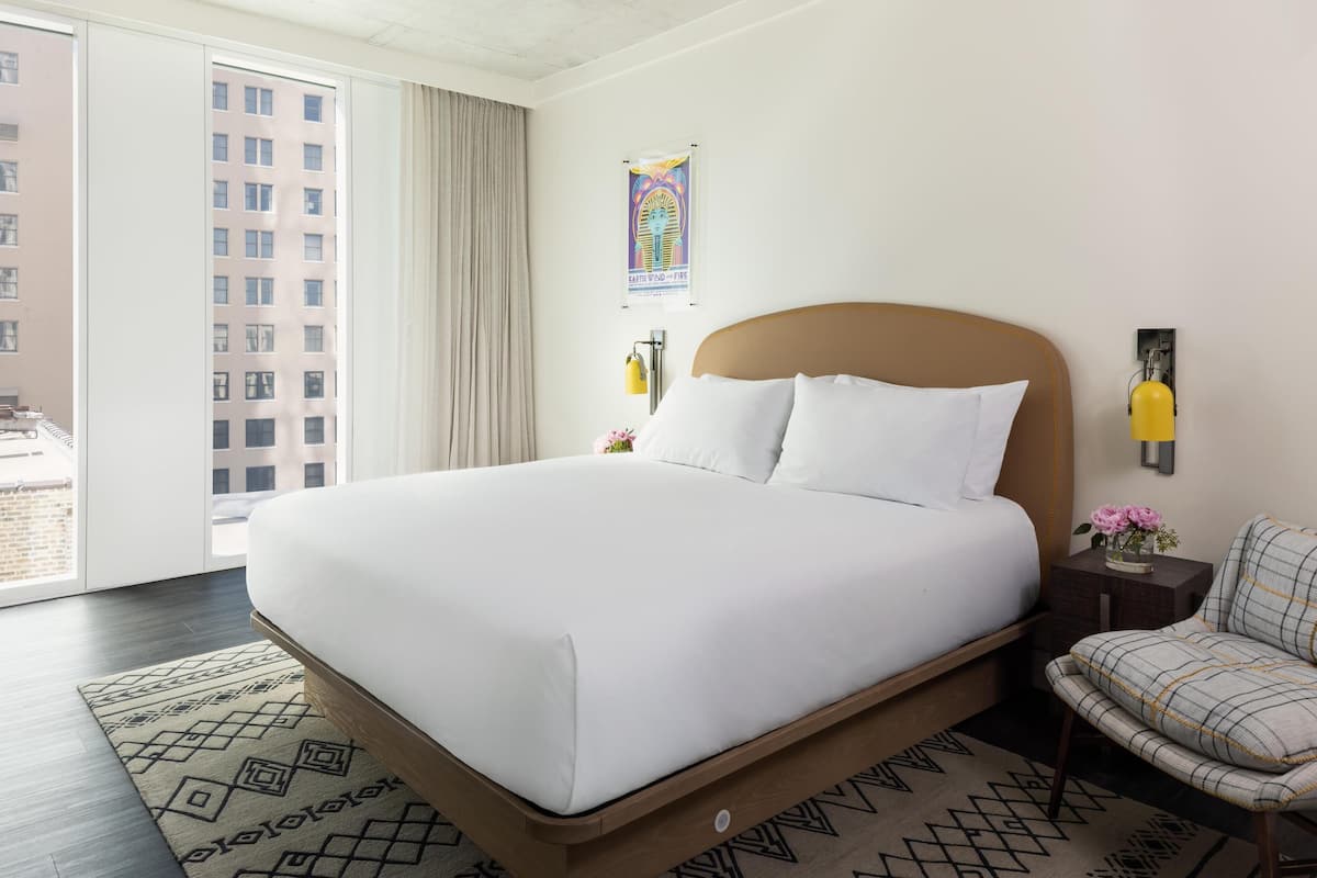 A hotel room with a large bed, a chair, a small table with a vase of flowers, a poster on the wall, and a window with curtains.