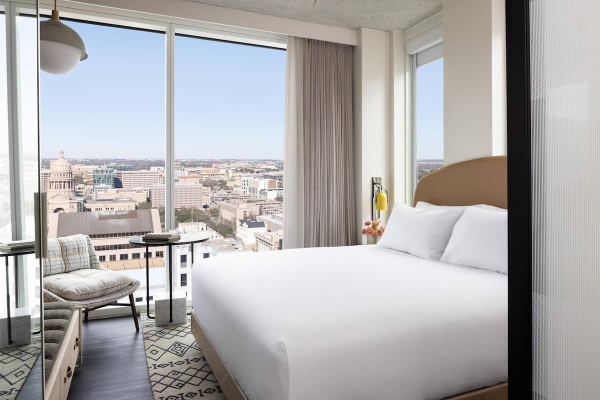 A hotel room with a large bed, a chair, a small table, and a view of a cityscape through large windows.