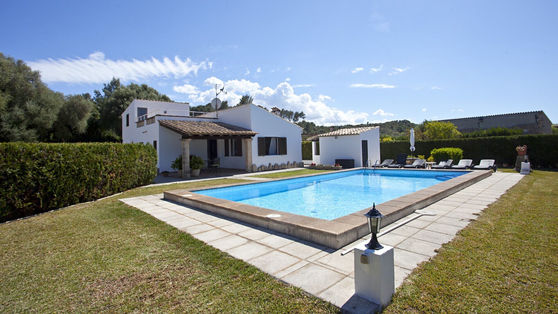 A rectangular swimming pool with a tiled deck, surrounded by a well-maintained lawn and a white building with a tiled roof.