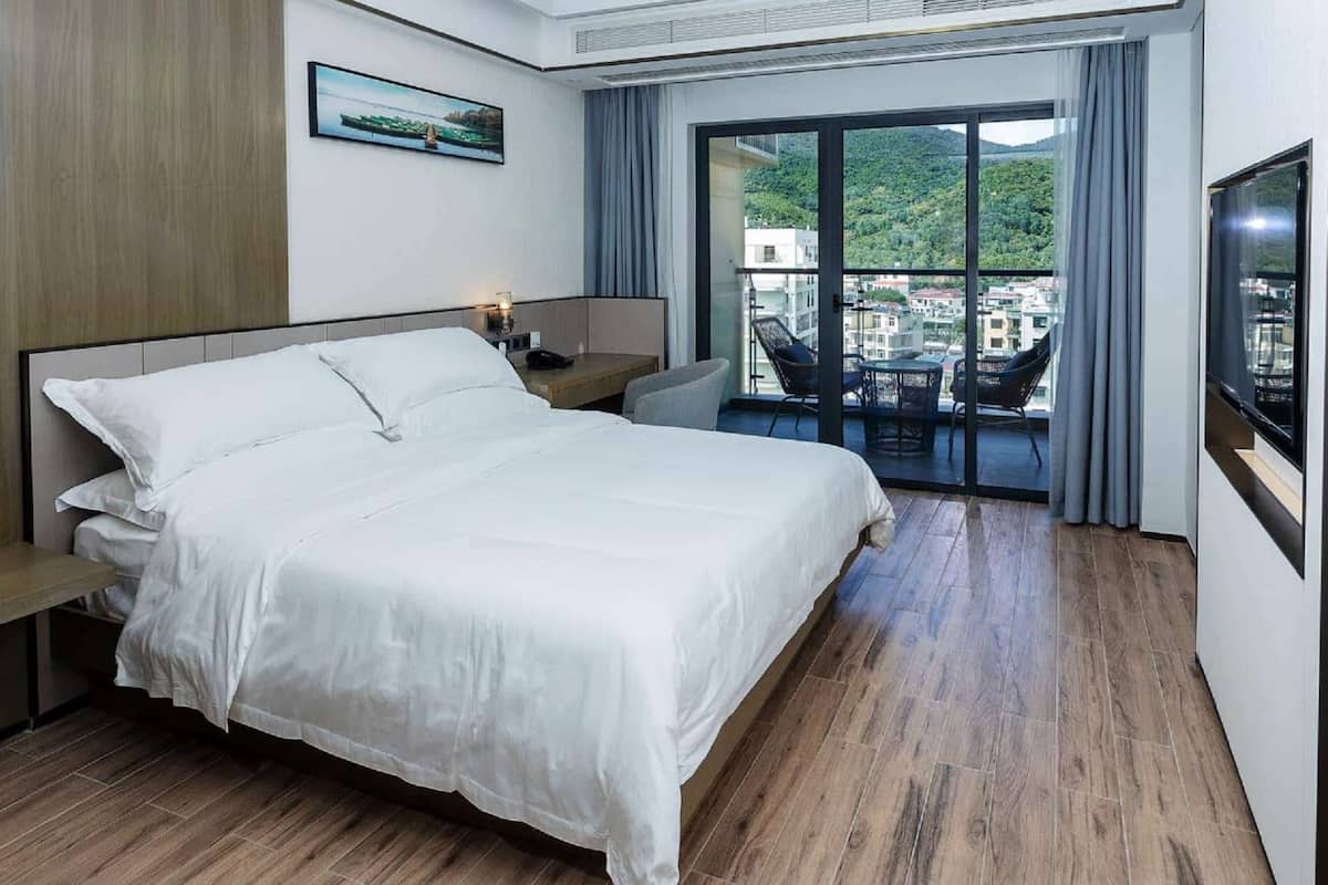 A hotel room with a large bed, a desk, a chair, and a balcony with a view of buildings and greenery.