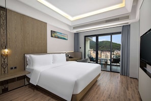 Quarto de hotel moderno com cama grande, escrivaninha, cadeira, televisão e varanda com vista para a cidade.
