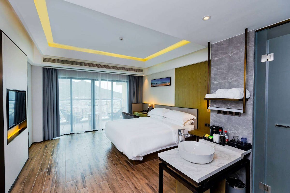 A hotel room with a bed, a bedside table with a bowl, a mirror, a TV, and a view of the city through large windows.