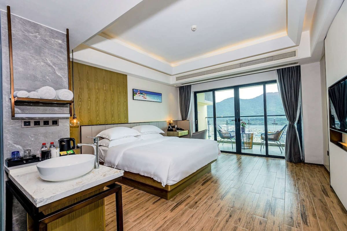 A hotel room with a large bed, a bathroom with a sink and mirror, a TV, and a balcony with a view of the sea.
