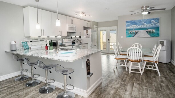 A modern kitchen with a central island, bar stools, and a dining area with white chairs. - Coastal Getaway - Steps from the Beach! (Cape Canaveral)