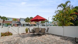 A patio with a table and chairs under a red umbrella, surrounded by a white fence and palm trees. - Coastal Getaway - Steps from the Beach! (Cape Canaveral)