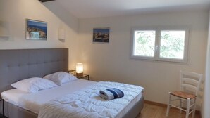 Un dormitorio con cama, mesita de noche con lámpara, una silla y dos ventanas.