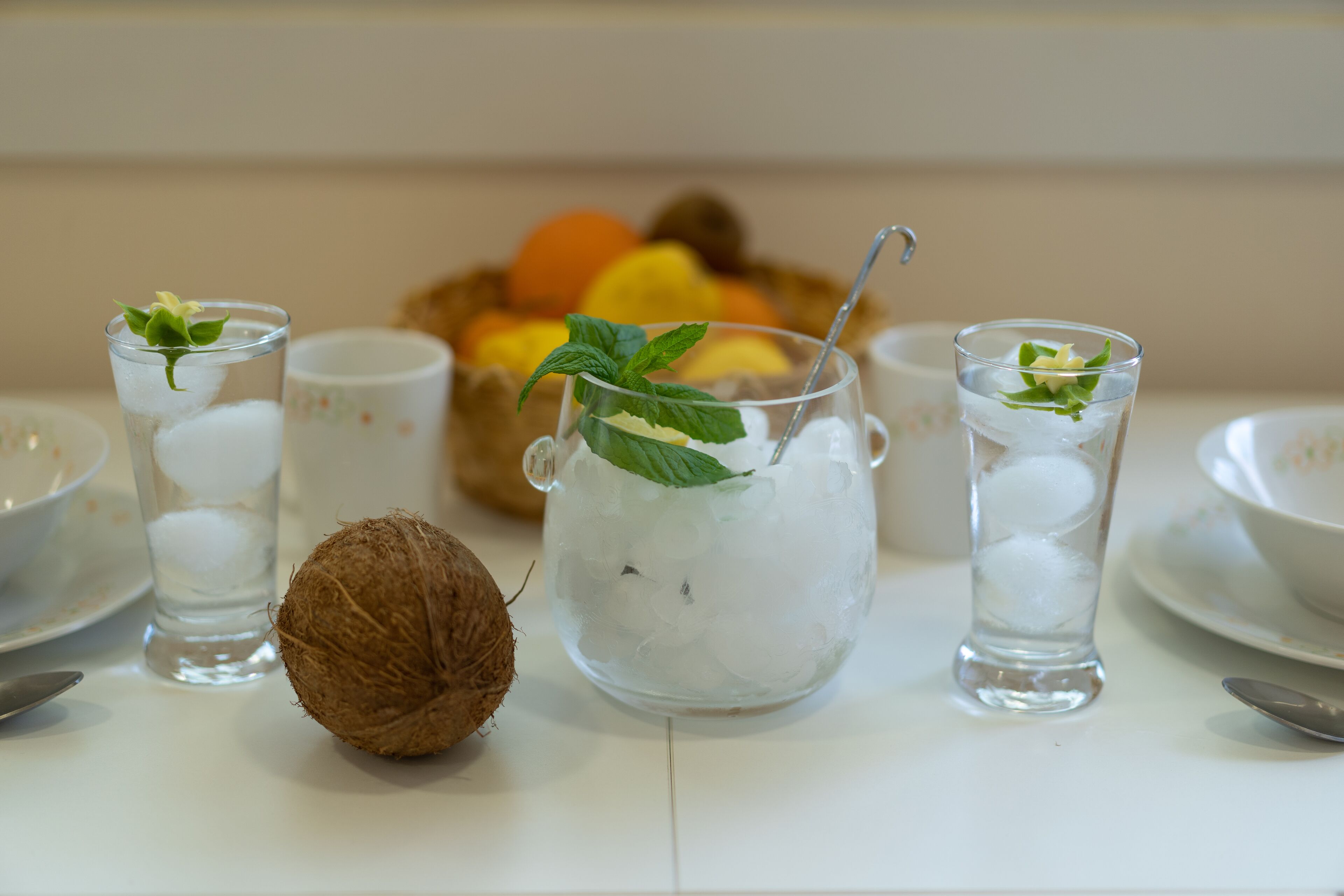 Een tafel met drie glazen verfrissende drankjes, een kokosnoot, en een fruitschaal op de achtergrond.