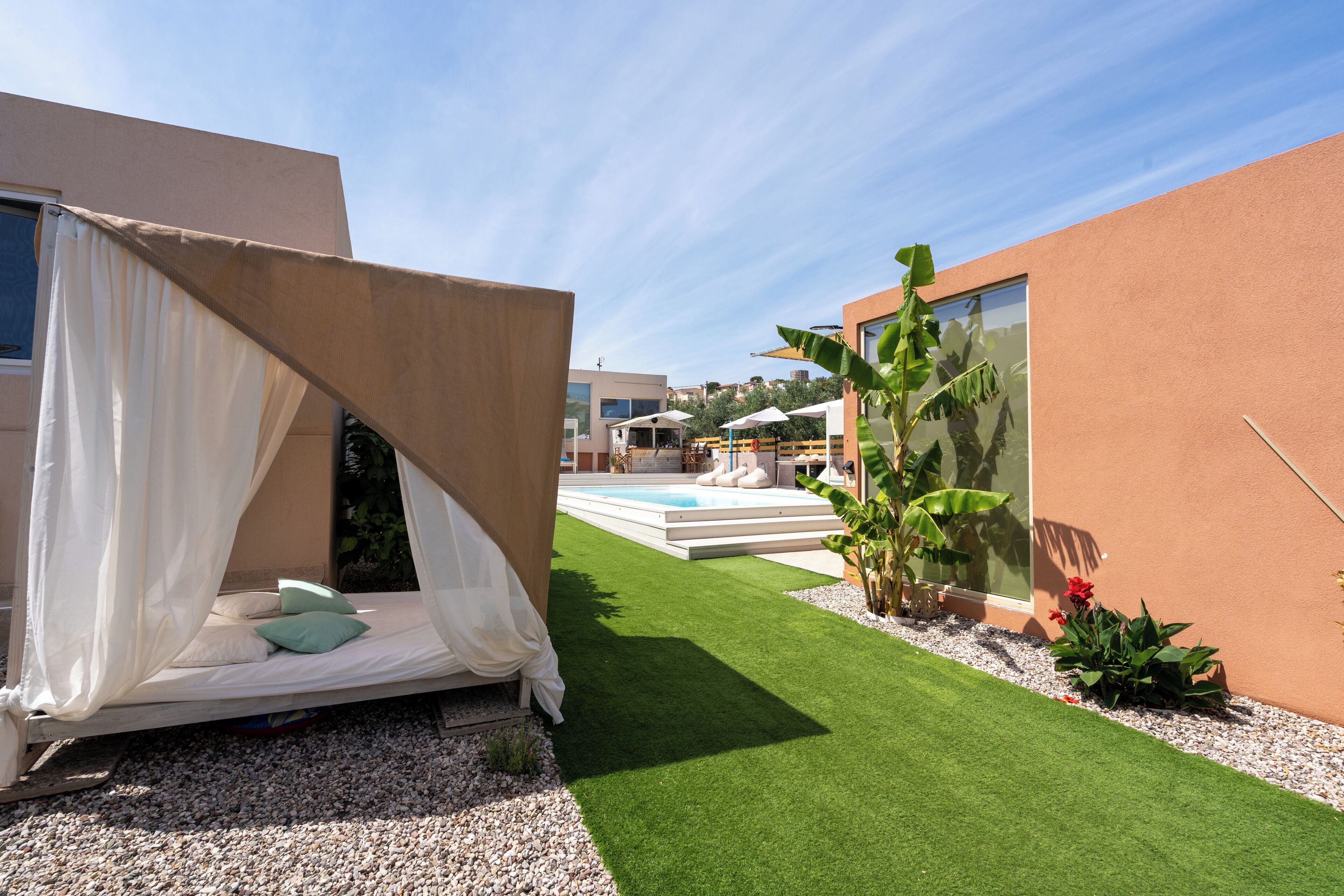 Un patio exterior moderno con cama, césped artificial, piscina y un edificio con amplios ventanales.