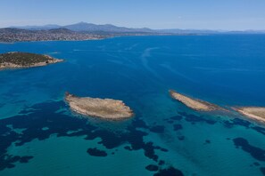 An aerial view of a coastal area with clear blue waters, rocky islands, and distant mountains. - Modular Bungalows With Heated Pool Artemis Greece (Spata-Artemida)