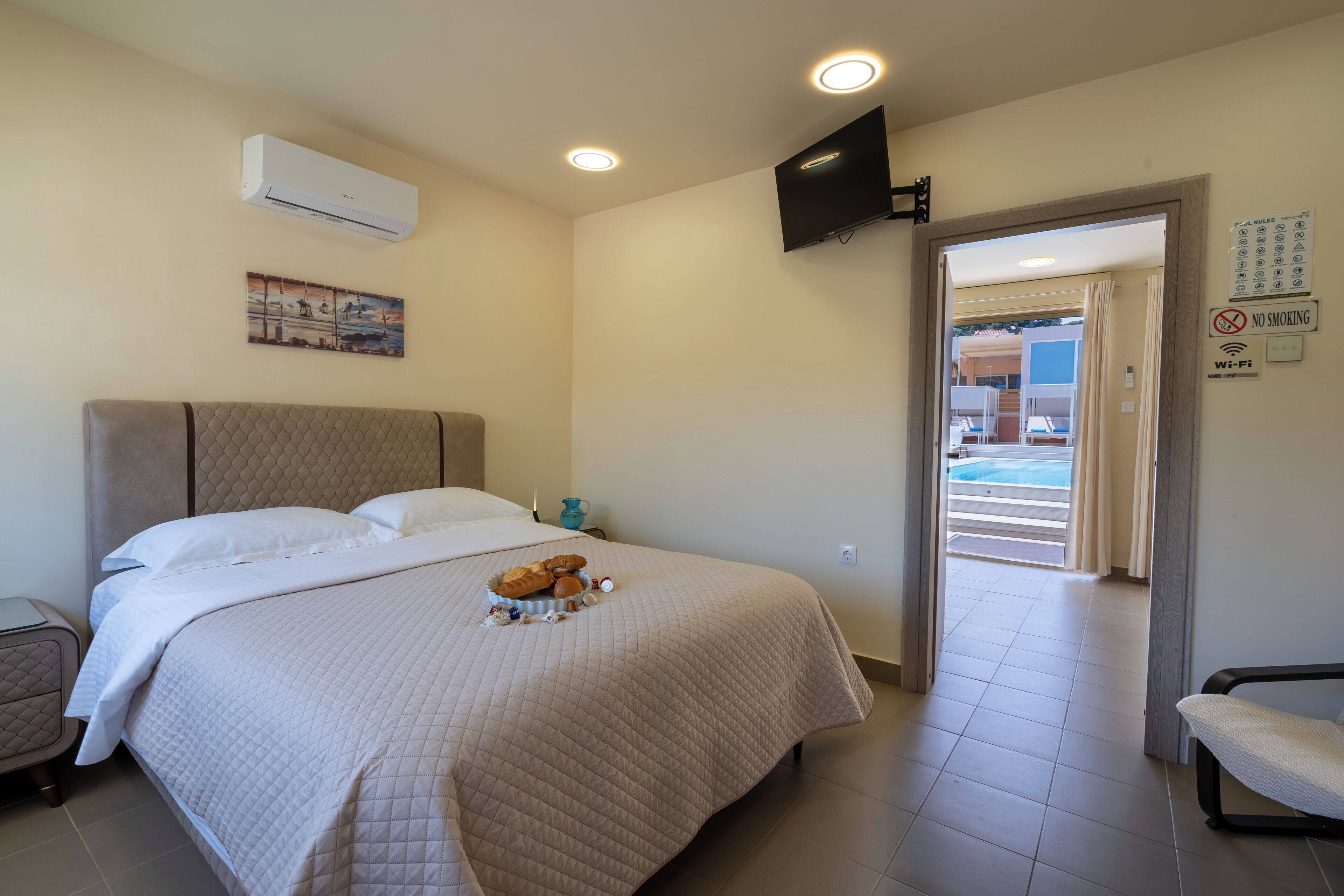 A hotel room with a bed, a flat-screen TV mounted on the wall, an air conditioning unit, a painting, and a door leading to a balcony.