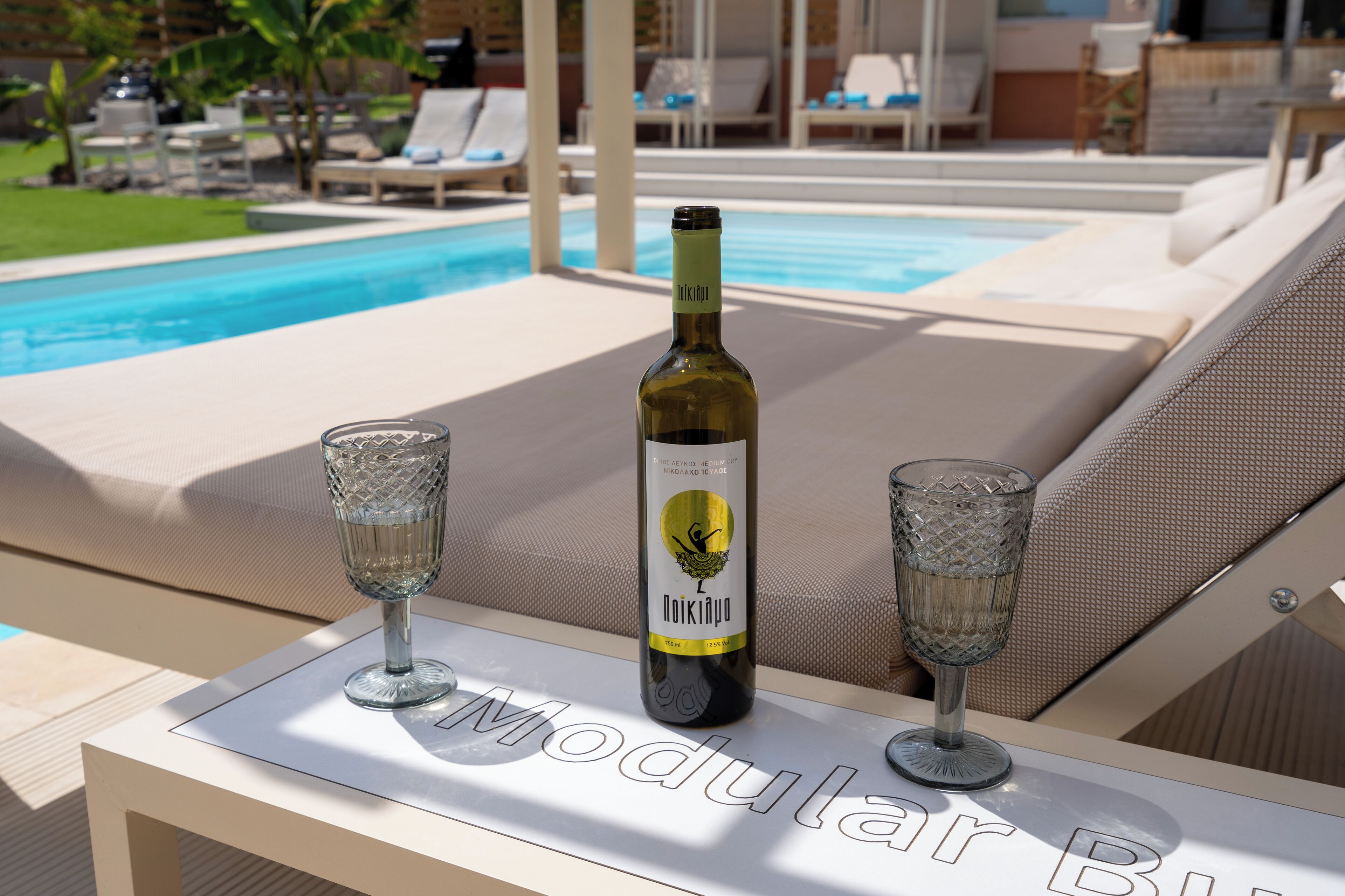 Una botella de vino Noikano y dos copas sobre una mesa cerca de una zona de descanso junto a la piscina.