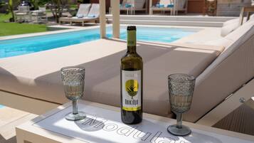 Una botella de vino Noikano y dos copas sobre una mesa cerca de una zona de descanso junto a la piscina.