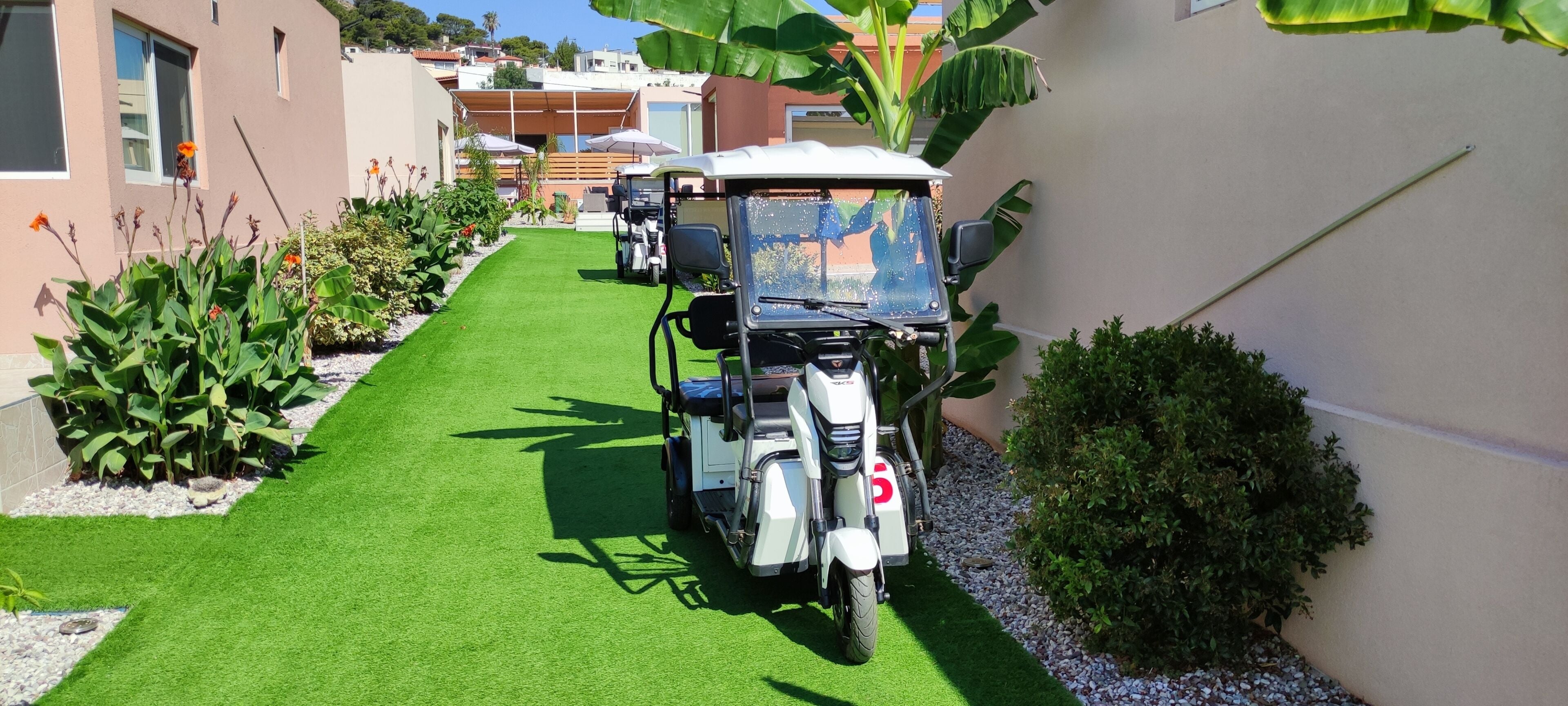 Un sendero estrecho con césped artificial verde, un carro de golf blanco y un edificio con fachada color crema.