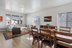 A spacious living room with a dining area, featuring a sofa, chairs, a wooden dining table, and large windows with blinds.