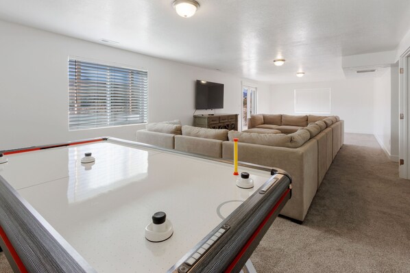 A room with a pool table, a beige sectional sofa, a flat-screen TV mounted on the wall, and a window with blinds.