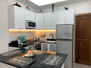 A modern kitchen with white cabinets, a stainless steel refrigerator, a microwave, a stove, and a coffee maker. There are two black countertops with decorative mats.