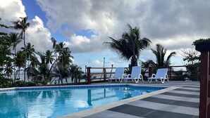 Une piscine bordée de carreaux, des chaises longues, des palmiers et un ciel nuageux.