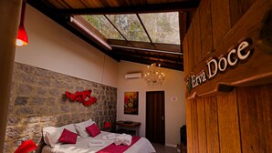 A bedroom with a stone wall, a bed with white and red bedding, a chandelier, and a wooden door with a sign. - Espaço Viverde Pousada (Itatiaia)