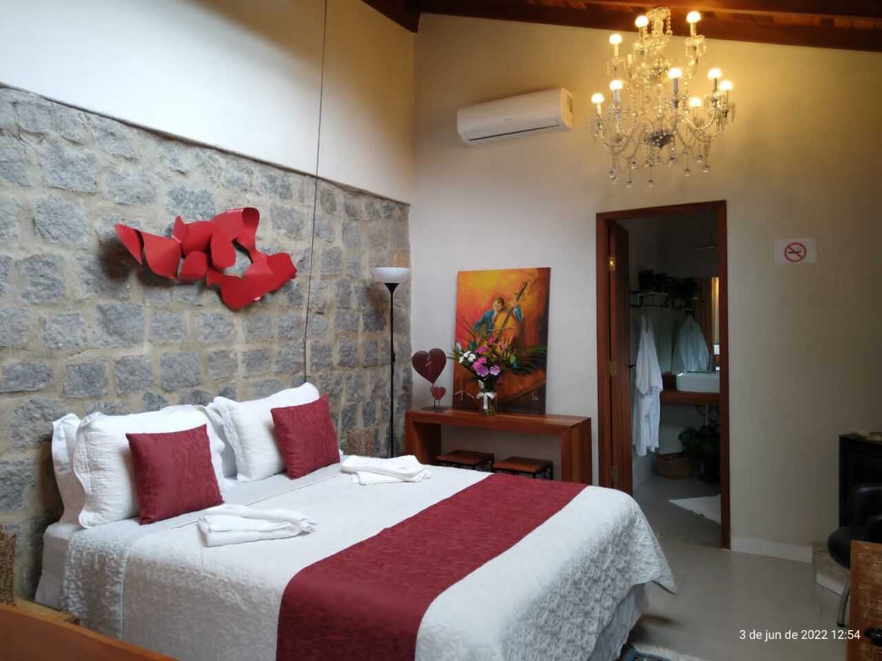A bedroom with a stone wall, a chandelier, a painting, and a bathroom visible through an open door.