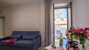 A modern hotel room with a dark blue sofa, a glass table with a book, glasses, a bottle, and a vase with flowers. - Neva-Elegant Suite with Balcony in Massa Lubrense center 10 minutes from Sorrento (Massa Lubrense)