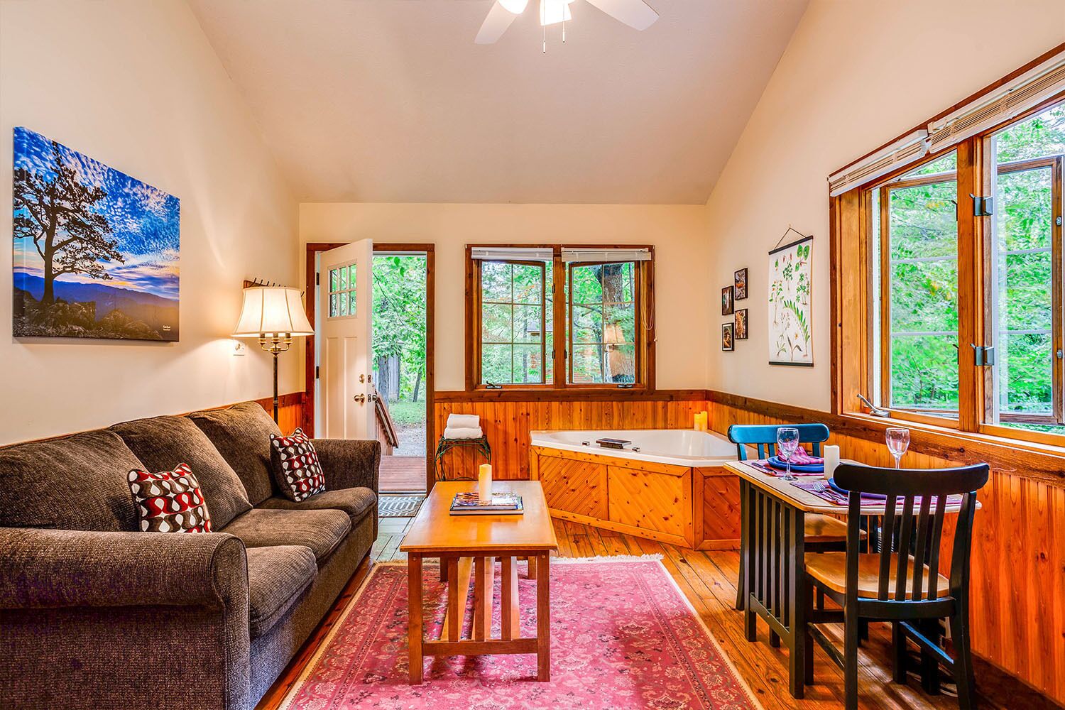 A cozy living room with a sofa, coffee table, and a dining area with a chair and table. There is a large window with a view of greenery outside.