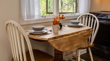 Una mesa de comedor de madera con dos sillas blancas, una ventana con cortinas blancas y vistas a árboles.