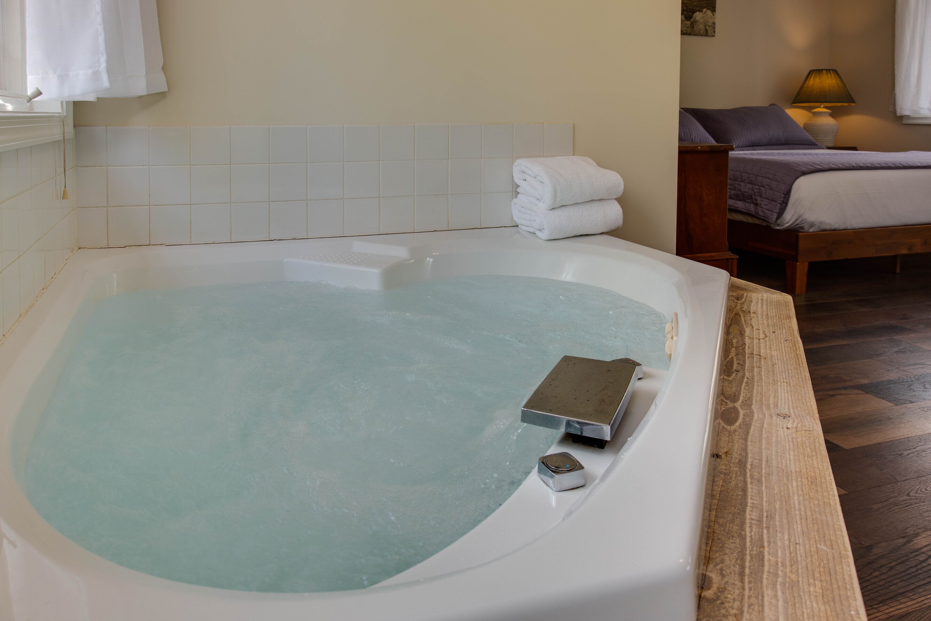 A room with a bathtub, a bed with a wooden headboard, a bedside table with a lamp, and a wall-mounted towel rack.