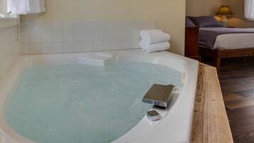 A room with a bathtub, a bed with a wooden headboard, a bedside table with a lamp, and a wall-mounted towel rack.