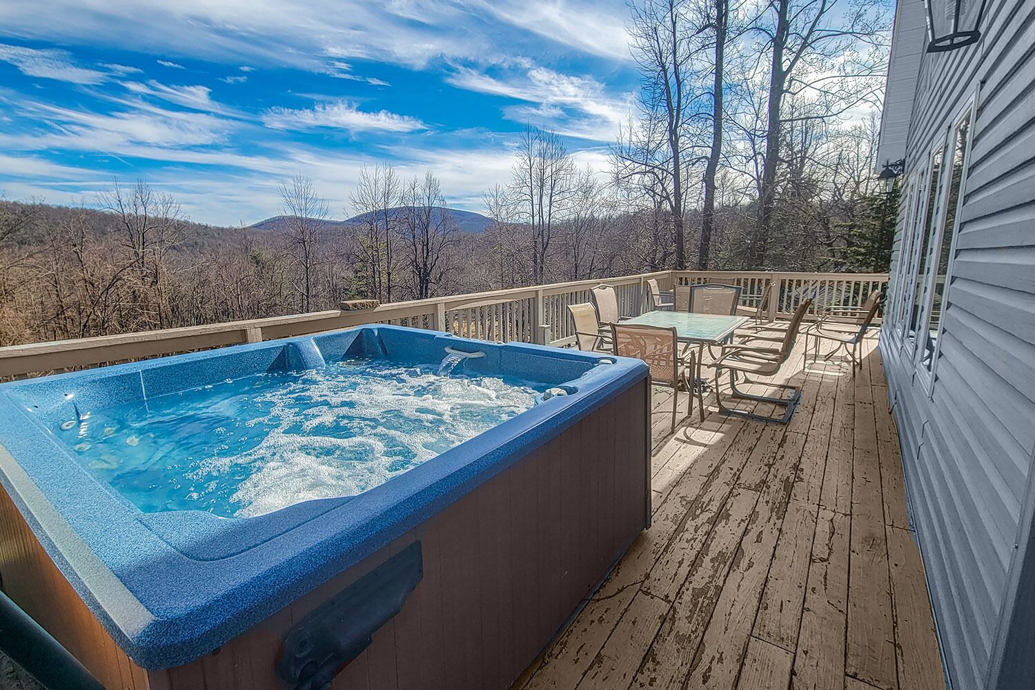 A hot tub on a wooden deck with a table and chairs, overlooking a mountainous landscape.