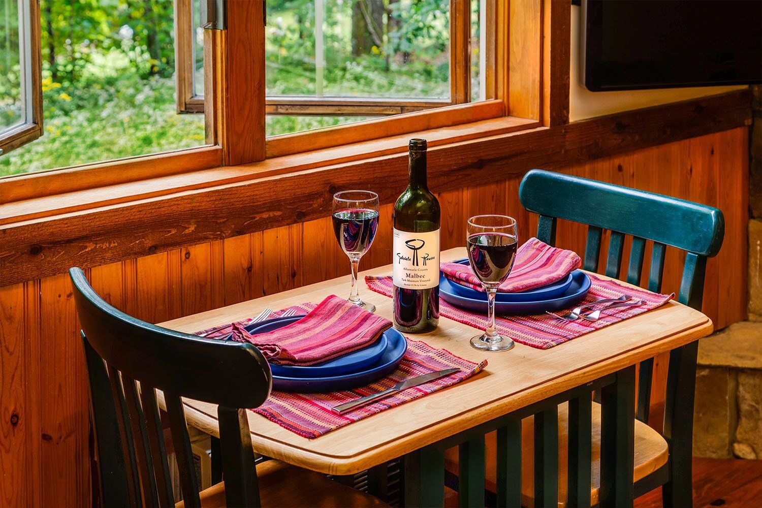 A wooden table with a bottle of wine, two glasses, and blue plates set on a bench with a view of greenery outside.