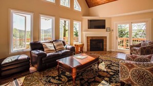 A spacious living room with a fireplace, leather sofa, patterned armchairs, and a large window overlooking a deck.