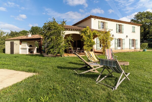 A house with a patio, a small structure with a chimney, and a lawn with two lounge chairs.