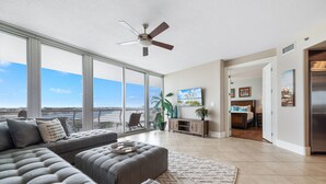 A spacious living room with a large gray sectional sofa, a ceiling fan, and a view of the ocean through floor-to-ceiling windows.
