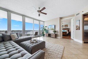 A spacious living room with a large gray sectional sofa, a ceiling fan, and a view of the ocean through floor-to-ceiling windows.