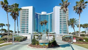 A modern hotel with a curved glass facade, palm trees, and a central sculpture.