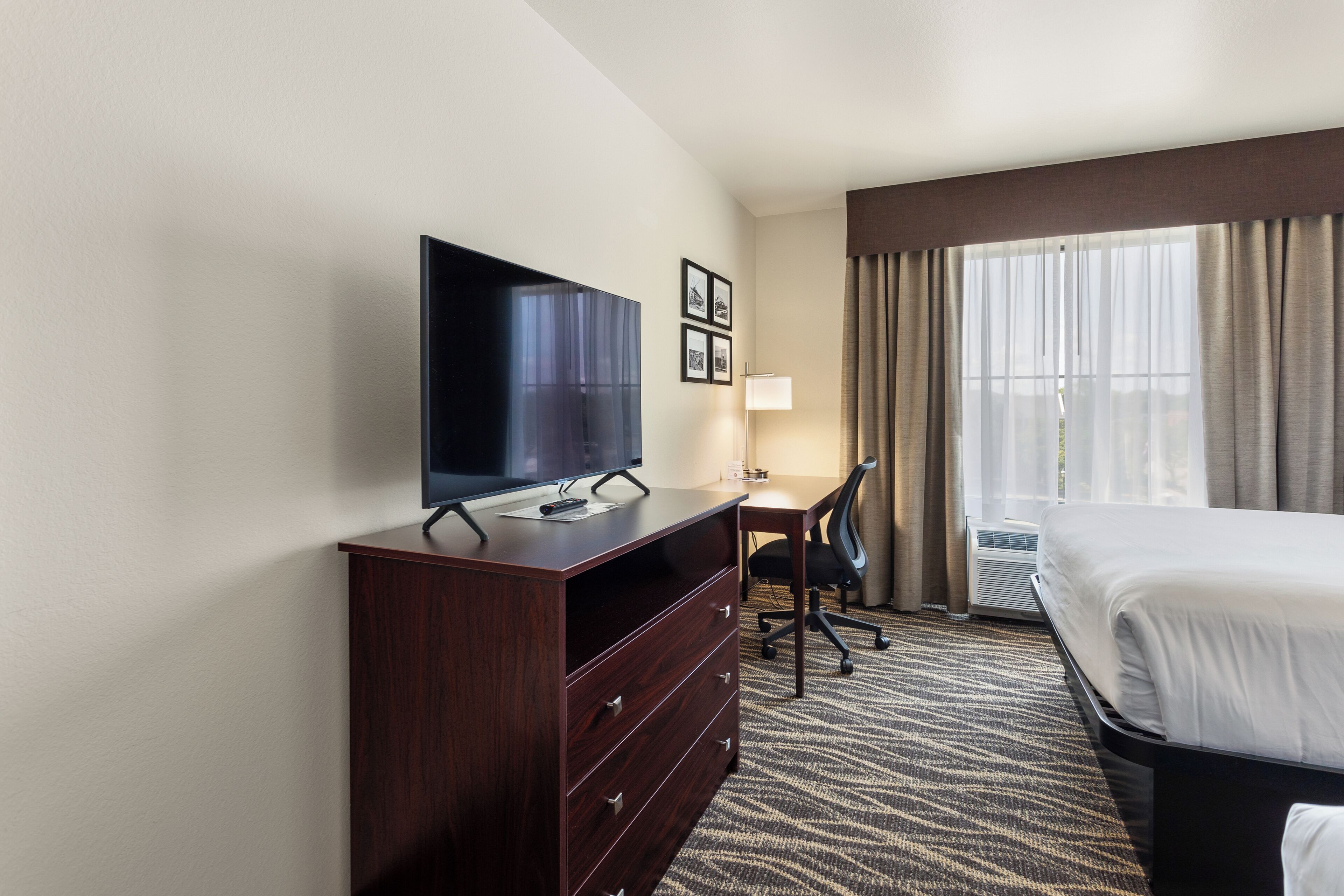 A hotel room with a bed, a TV on a wooden dresser, a desk with a chair, and a large window with curtains.