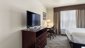 A hotel room with a bed, a TV on a wooden dresser, a desk with a chair, and a large window with curtains.