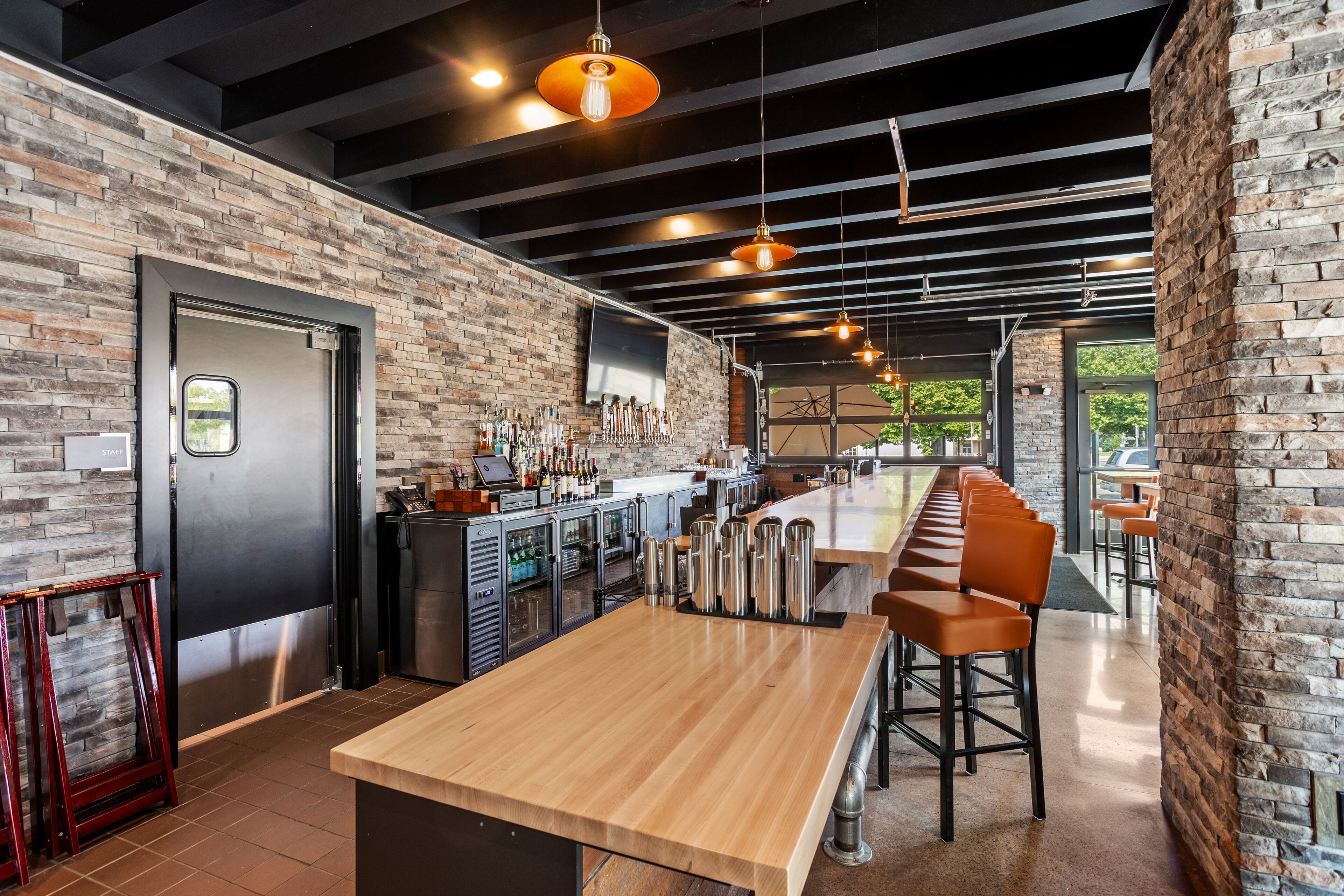 A modern bar area with a long wooden counter, bar stools, and a brick wall.