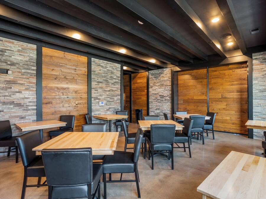 Un restaurante moderno con mesas de madera y sillas negras, paredes de ladrillo visto y un techo alto con iluminación empotrada.