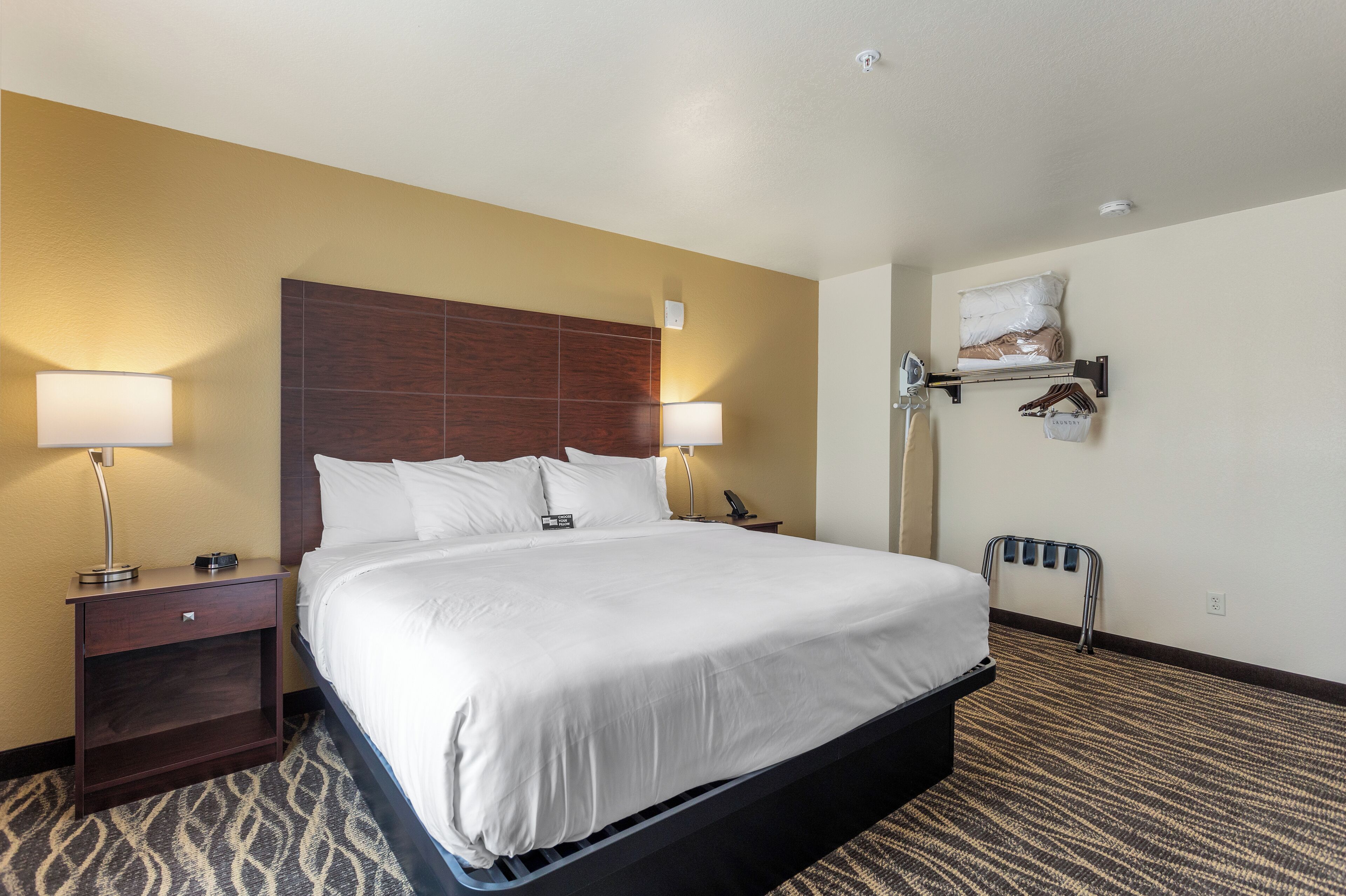 A hotel room with a large bed, two bedside tables with lamps, a wall-mounted shelf with bags, and a wheelchair-accessible bench.