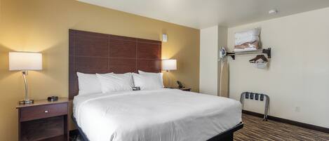 A hotel room with a large bed, two bedside tables with lamps, a wall-mounted shelf with bags, and a wheelchair-accessible bench.